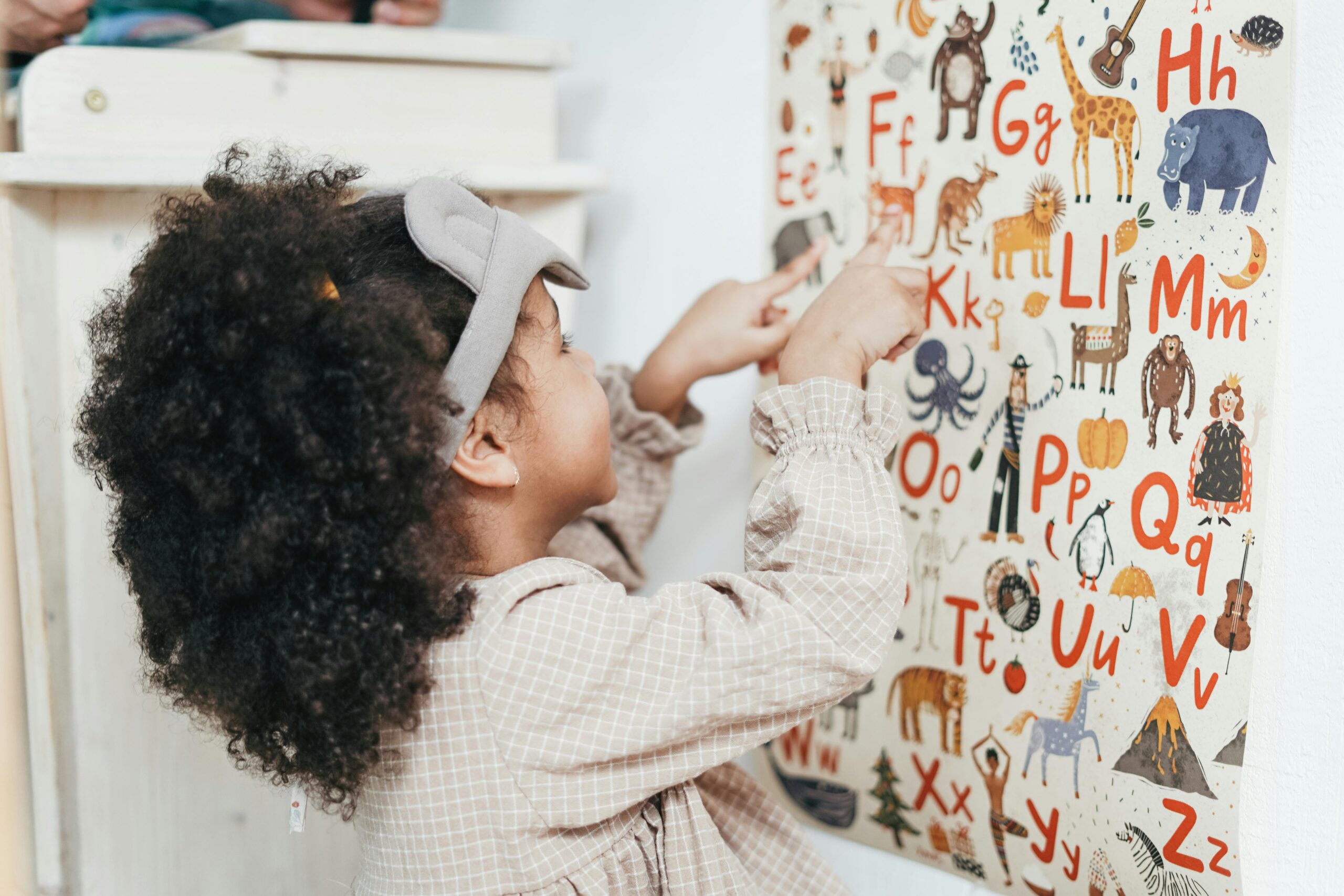 Child with curly hair points to an animal alphabet poster indoors, learning letters and animals.