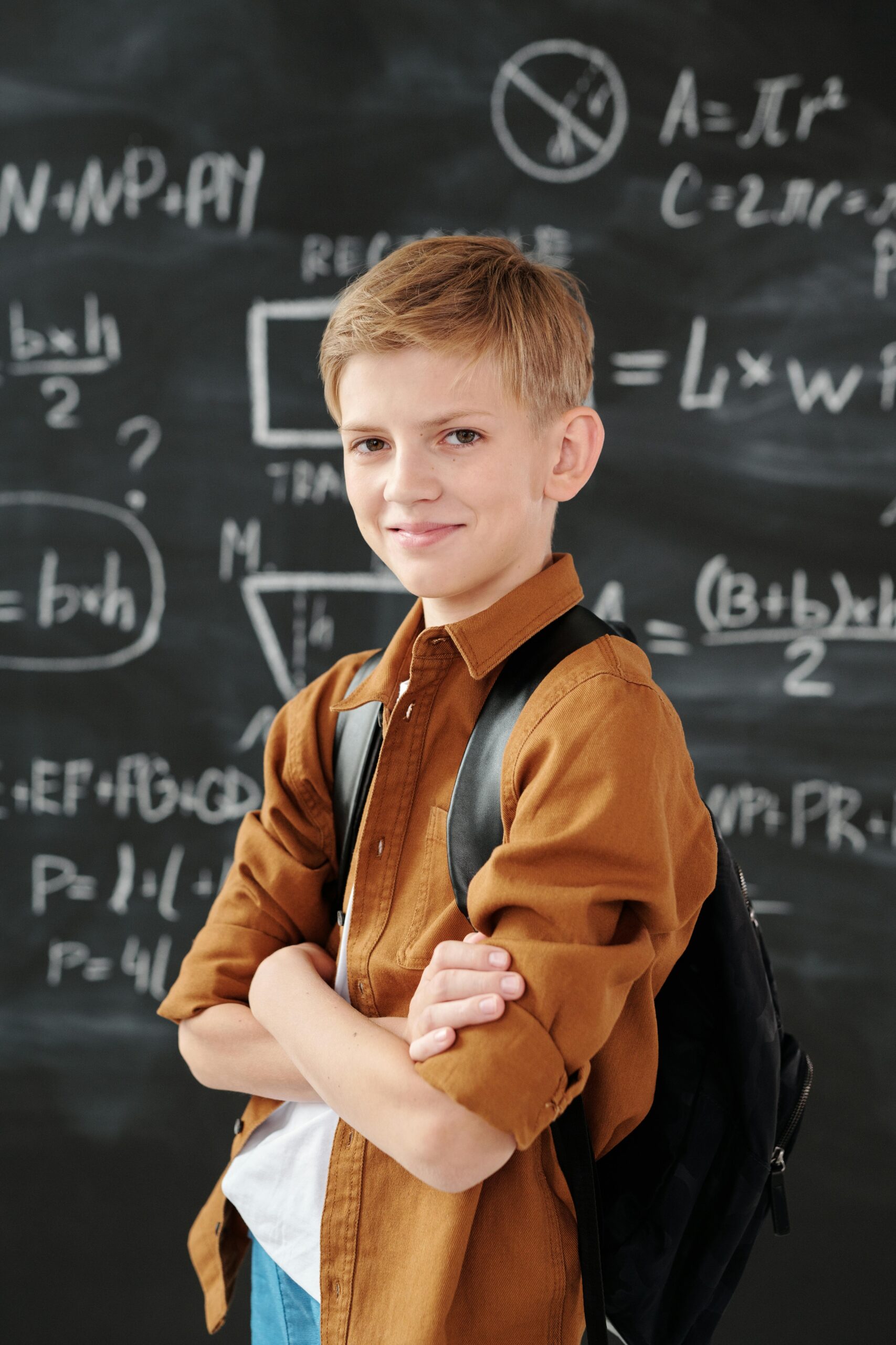 Smiling elementary student standing in classroom with math equations on blackboard.